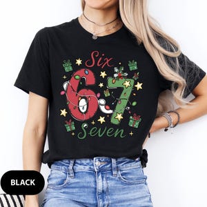 May include: Black t-shirt with a festive design featuring the numbers "6" and "7" in red and green, adorned with Christmas lights, stars, and gift boxes. The words "Six" and "Seven" are also displayed.