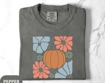 May include: A gray Comfort Colors t-shirt with a retro floral and pumpkin design. The design features an orange pumpkin, blue and coral flowers, and green accents. The shirt has a crew neck and a tag that reads "Comfort Colors".