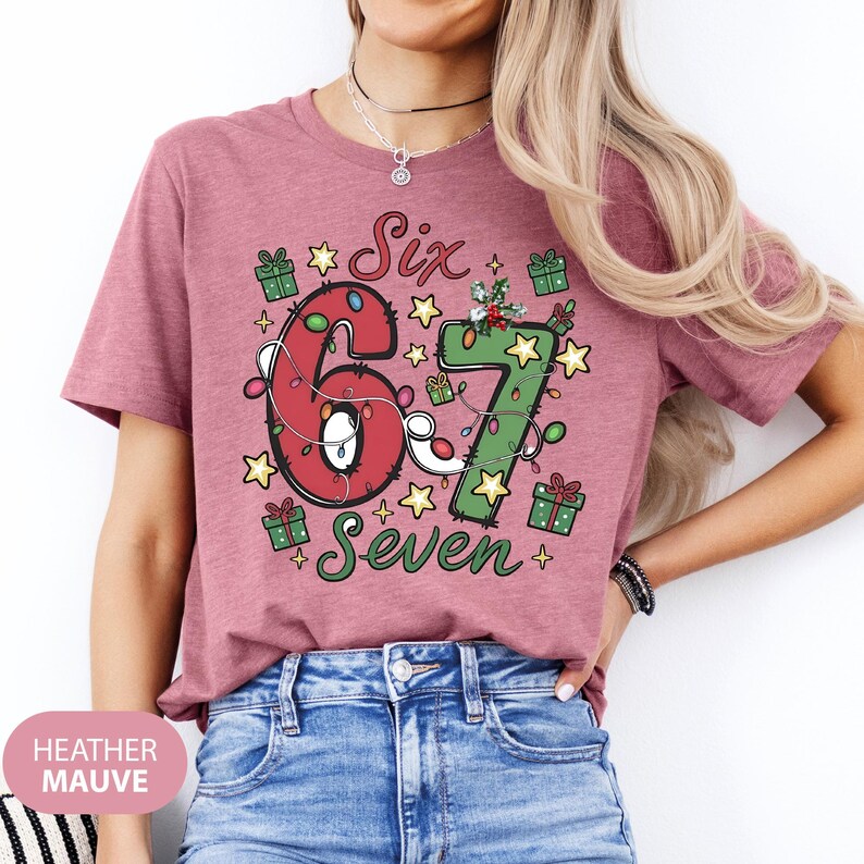 May include: Heather mauve t-shirt with a festive design. The graphic features the numbers "6" and "7" decorated with Christmas lights, surrounded by stars and gift boxes. The words "Six" and "Seven" are also included.