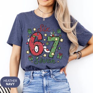 May include: Heather navy blue t-shirt with a festive design. The shirt features the words "Six" and "Seven" with the numbers 6 and 7 decorated with Christmas lights and stars. Small gift boxes are scattered around the numbers.