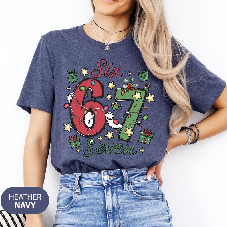 May include: Heather navy blue t-shirt with a festive design. The shirt features the words "Six" and "Seven" with the numbers 6 and 7 decorated with Christmas lights and stars. Small gift boxes are scattered around the numbers.