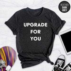 May include: Dark heather grey t-shirt with the text "UPGRADE FOR YOU" in white. The shirt is a crew neck and has rolled-up sleeves. A black circle with the words "HEATHER DARK GREY" is in the upper right corner.