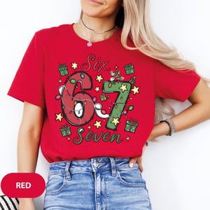 May include: A red t-shirt with the words "Six Seven" in a festive design. The numbers are decorated with Christmas lights and surrounded by stars and gift boxes. The shirt is a solid red color.