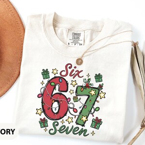 May include: Ivory-colored t-shirt with the numbers "6" and "7" decorated with Christmas lights and the words "Six" and "Seven". The shirt has a crew neck and short sleeves. A gold necklace is draped on the shirt.