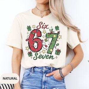 May include: Cream-colored t-shirt with a festive Christmas design. The graphic features the numbers "6" and "7" decorated with lights, surrounded by stars and gift boxes. The words "Six" and "Seven" are also included in the design.