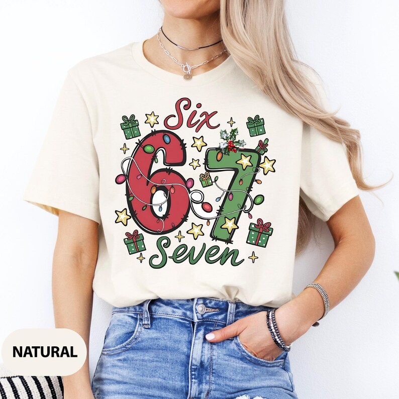 May include: Cream-colored t-shirt with a festive Christmas design. The graphic features the numbers "6" and "7" decorated with lights, surrounded by stars and gift boxes. The words "Six" and "Seven" are also included in the design.