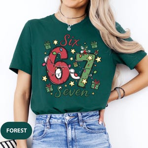 May include: Forest green t-shirt with a festive design. The graphic features the numbers "6" and "7" decorated with Christmas lights, stars, and gift boxes. The word "Six" and "Seven" are also included in the design.