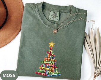 May include: A moss green t-shirt with a crayon Christmas tree design. The tree is made of colorful crayons and topped with a gold star. A gold necklace is draped across the shirt.