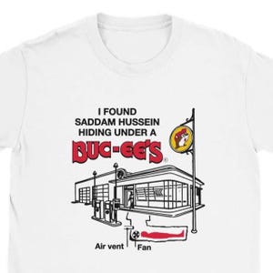 Könnte beinhalten: Weißes T-Shirt mit dem Aufdruck "I FOUND SADDAM HUSSEIN HIDING UNDER A BUC-EE'S". Die Grafik zeigt eine Cartoon-Tankstelle und ein Schild mit einem Biber-Logo. Der Text ist in Schwarz und Rot gehalten.