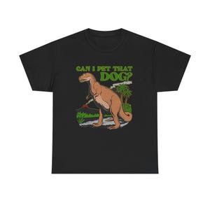 Op de afbeelding: Een zwart t-shirt met korte mouwen en een afbeelding van een bruine dinosaurus in een prehistorisch landschap met een vulkaan en palmbomen. De tekst boven de dinosaurus luidt CAN I PET THAT DOG? in dikke groene hoofdletters.