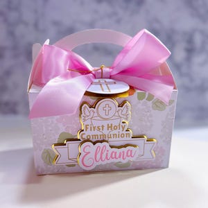 May include: A small gift box with a pink satin bow and handle. The box is white with a floral pattern and gold accents. Text on the box reads "First Holy Communion" and the name "Elliana".
