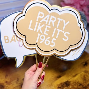 Puede incluir: Accesorios de fotomatón con detalles dorados. Un accesorio tiene forma de nube con el texto "PARTY LIKE IT'S 1965". Otro es un bocadillo con la palabra "BA". Los accesorios son sostenidos por una persona con esmalte de uñas rojo.