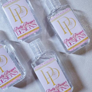 Puede incluir: Cuatro botellas pequeñas y transparentes de desinfectante de manos con etiquetas blancas. Las etiquetas presentan un logotipo "PP" dorado y el texto "Pretty Prosper DESIGNS" en rosa y dorado. Las botellas están sobre una superficie blanca brillante.