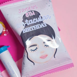 May include: A pink and white favor bag with a girl's face on it. The bag says "Jayden's 8th Spa-tacular Birthday".