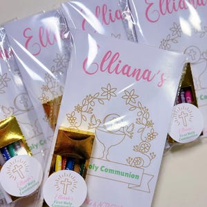 May include: Several First Holy Communion favors are displayed. Each favor includes a booklet with the name "Elliana's" in pink script, a gold-colored crayon box, and a sticker with a cross. The booklets have floral and dove designs.