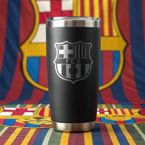 Pode incluir: Um copo preto de aço inoxidável com aro e base prateados. O copo apresenta um brasão prateado do FC Barcelona. O fundo inclui as cores e o logótipo da equipa do FC Barcelona.