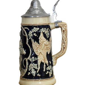 May include: A ceramic beer stein with a dark blue background and cream-colored relief carvings of a bird and foliage. The stein has a silver metal lid and handle, and a cream-colored base. The handle is decorated with relief carvings.