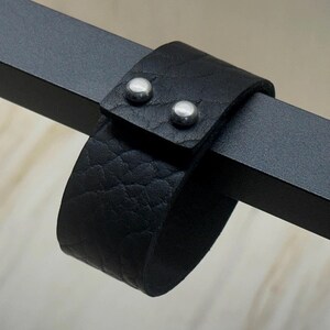 Op de afbeelding: Een zwarte leren armband met twee zilveren klinknagels. De armband is te zien op een zwart metalen oppervlak.