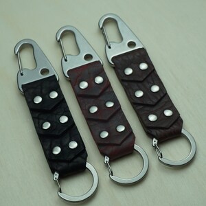 May include: Three leather keychains with metal fixings. The keychains have a dark brown, burgundy, and black leather strap with silver rivets and a silver key ring. The keychains are designed for keys.