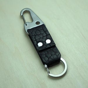 May include: A black leather keychain with a silver carabiner clip and a split ring. The leather has a textured appearance and is secured with two silver rivets. The keychain is designed for attaching keys or other small items.