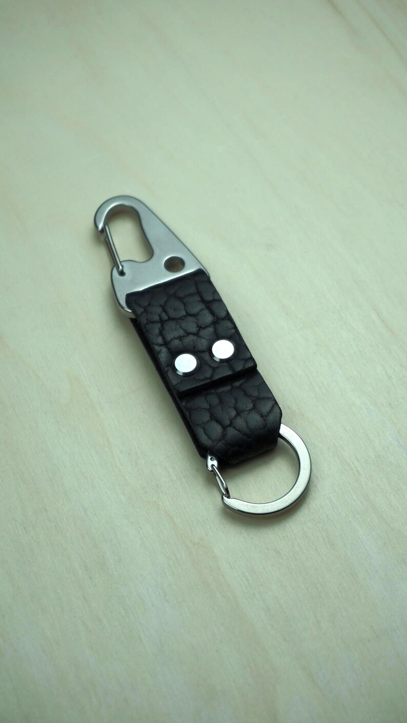 May include: A black leather keychain with a silver carabiner clip and a split ring. The leather has a textured appearance and is secured with two silver rivets. The keychain is designed for attaching keys or other small items.