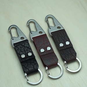 May include: Three leather keychains with silver-tone hardware. The keychains feature textured leather straps in black, burgundy, and dark brown. Each keychain has a metal clasp and a split ring.