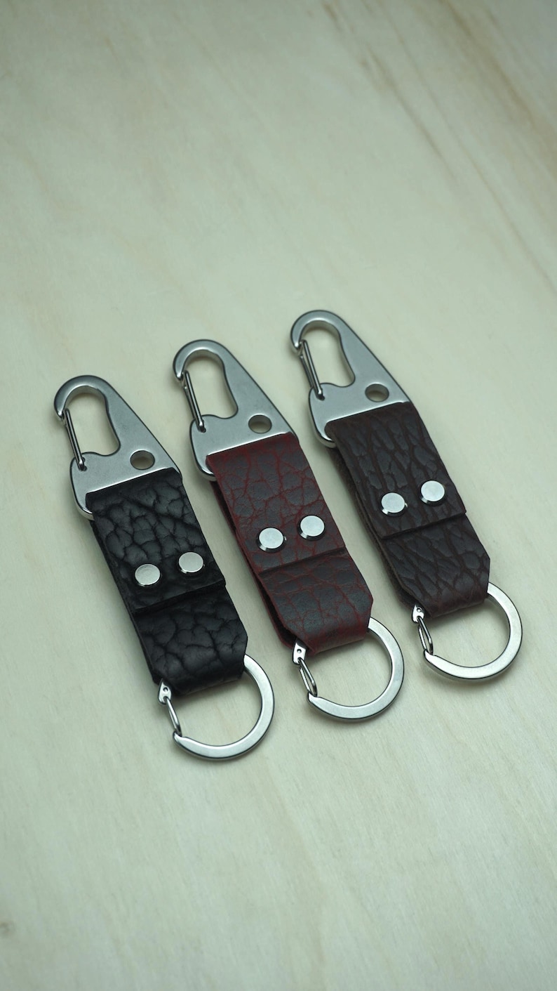 May include: Three leather keychains with silver-tone hardware. The keychains feature textured leather straps in black, burgundy, and dark brown. Each keychain has a metal clasp and a split ring.