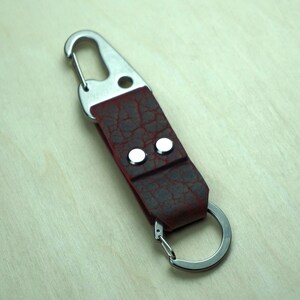 May include: A burgundy leather keychain with a silver-colored metal clasp and key ring. The leather has a textured, cracked appearance and is secured with two silver rivets. The keychain is designed for attaching keys or other small items.