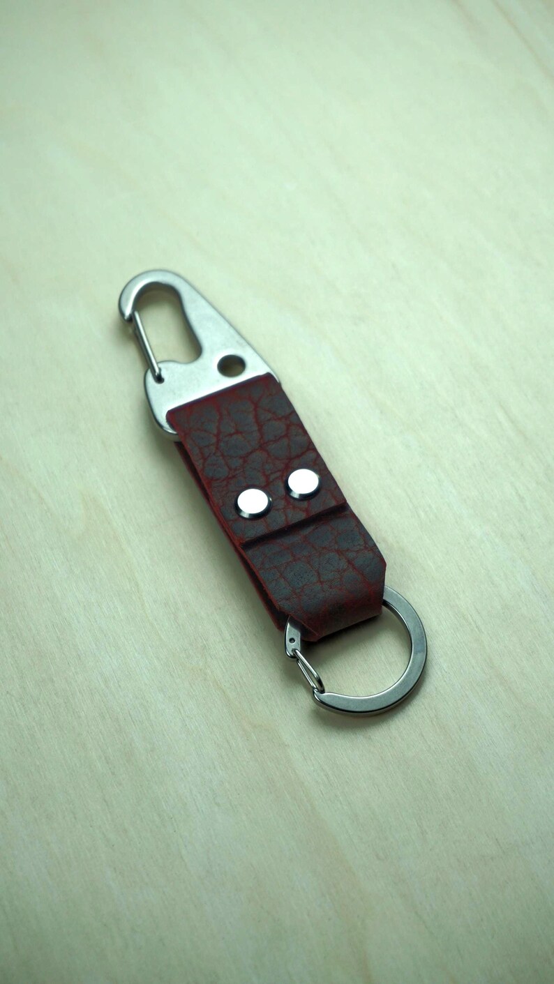 May include: A burgundy leather keychain with a silver-colored metal clasp and key ring. The leather has a textured, cracked appearance and is secured with two silver rivets. The keychain is designed for attaching keys or other small items.