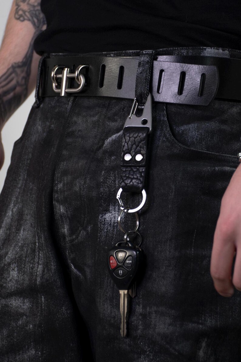 May include: Black leather belt with a silver buckle and a key holder attached. The key holder has a textured leather strap and a metal clip, holding a car key. The belt is worn with black denim jeans.