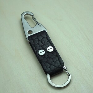 May include: A black leather keychain with a textured surface, featuring a silver-colored carabiner clip and a split ring. The leather strap is secured with two silver rivets.