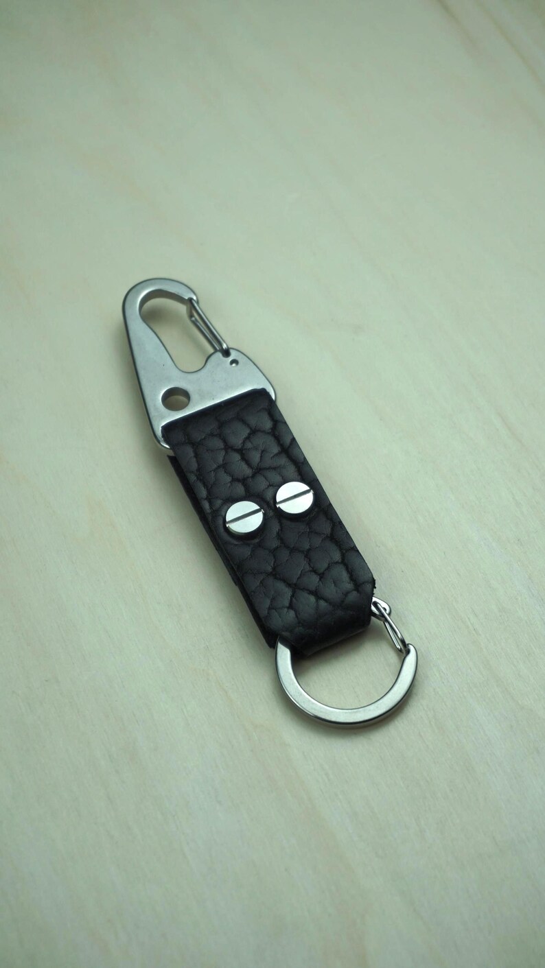 May include: A black leather keychain with a textured surface, featuring a silver-colored carabiner clip and a split ring. The leather strap is secured with two silver rivets.