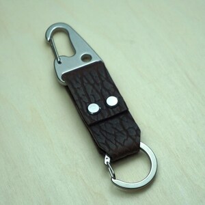 May include: A brown leather keychain with a silver carabiner clip and a split ring. The leather has a textured appearance and is secured with two silver rivets. The keychain is designed for attaching keys or other small items.