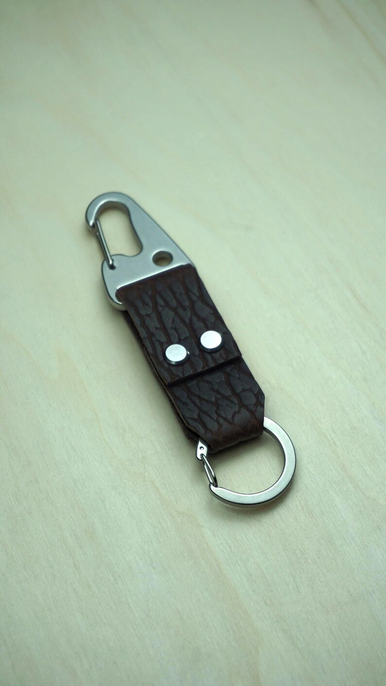 May include: A brown leather keychain with a silver carabiner clip and a split ring. The leather has a textured appearance and is secured with two silver rivets. The keychain is designed for attaching keys or other small items.