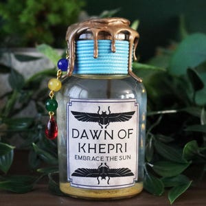 May include: A clear glass bottle with a gold-colored top and a blue string around the neck. The label reads "DAWN OF KHEPRI EMBRACE THE SUN" with a winged scarab design. A beaded charm hangs from the bottle.