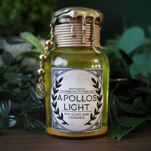 May include: Apothecary bottle with a gold-colored top and a label that reads "Apollos Light". The bottle is filled with a yellow liquid and is decorated with a gold charm. The bottle is surrounded by green foliage.