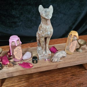 Puede incluir: Un altar de madera con una estatua de gato tallada, dos botellas con sellos de cera rosa y amarilla, un cristal de cuarzo rosa y dos dados negros con runas. Las botellas están etiquetadas como "Aphrodite's Kiss" y "Moon & Earth".