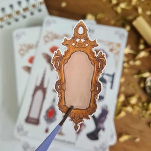 May include: A sticker featuring a detailed, ornate gold-colored mirror frame with a blank center. The sticker is being held by a pair of blue tweezers, with other stickers and a notebook in the background. The design has a vintage or fantasy theme.