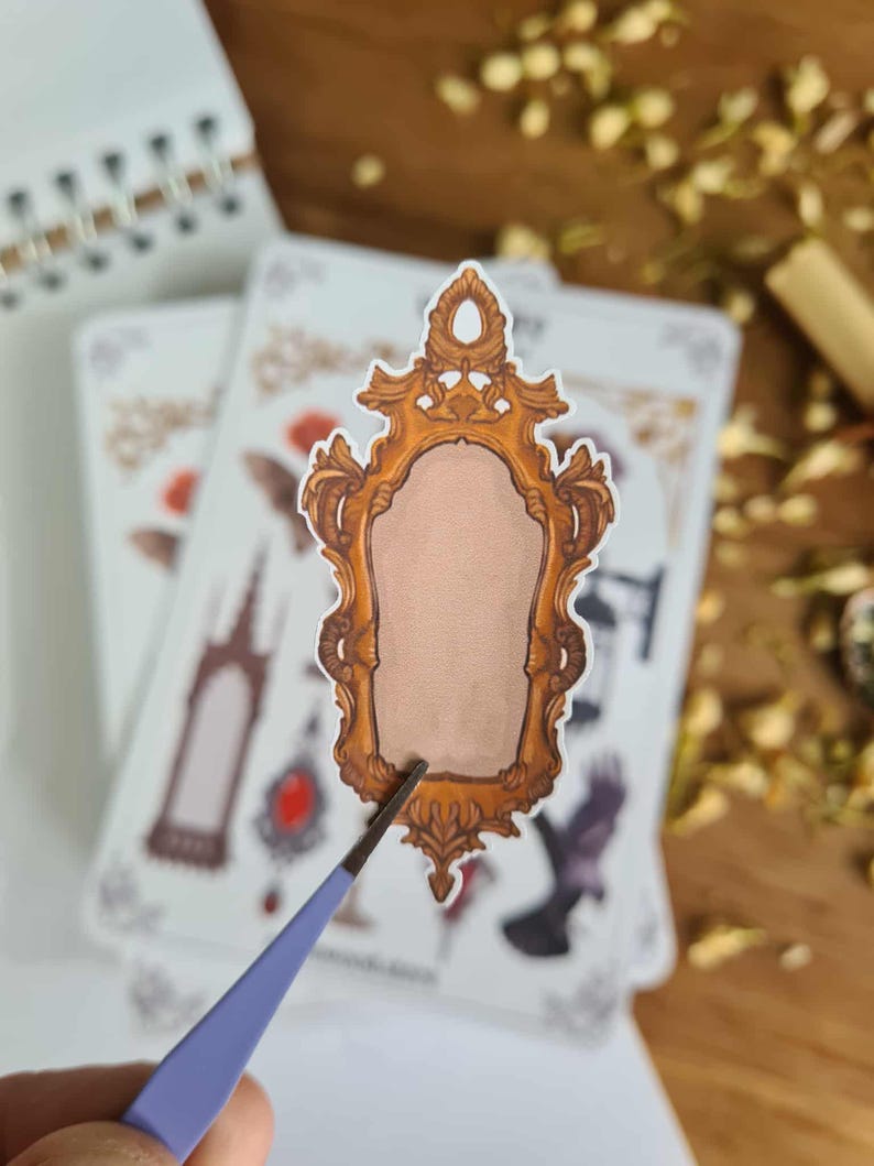 May include: A sticker featuring a detailed, ornate gold-colored mirror frame with a blank center. The sticker is being held by a pair of blue tweezers, with other stickers and a notebook in the background. The design has a vintage or fantasy theme.