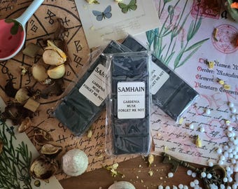 May include: Several black wax melt bars in clear packaging are displayed, labeled "SAMHAIN GARDENIA MUSK FORGET ME NOT." The image includes dried flowers, butterflies, and a decorative spoon, creating a rustic, botanical theme.