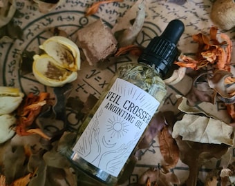 Può includere: Una bottiglia di vetro trasparente di "Veil Crosser Anointing Oil" con un contagocce nero. La bottiglia è circondata da foglie secche e elementi botanici su una superficie simile alla pergamena. L'etichetta presenta un sole e mani.