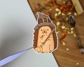 Könnte beinhalten: Ein skurriler Aufkleber mit einem Cartoon-Igel, der als Hexe verkleidet ist. Der Igel trägt einen braunen Hexenhut mit goldener Schnalle und hält einen Besen. Der Aufkleber hat einen hellbraunen Körper, braune Stacheln und ein lächelndes Gesicht.
