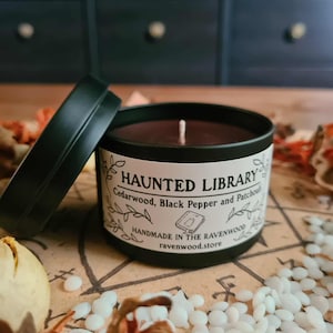 May include: A black tin candle with the text "HAUNTED LIBRARY" and scent notes. The candle is open, revealing a dark brown wax and a white wick. The label also includes "Cedarwood, Black Pepper and Patchouli".