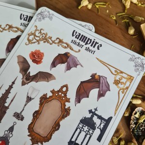 May include: A sheet of vampire-themed stickers featuring bats, a rose, a mirror, and other gothic elements. The stickers are on a white background with the words "vampire sticker sheet" printed on the top.