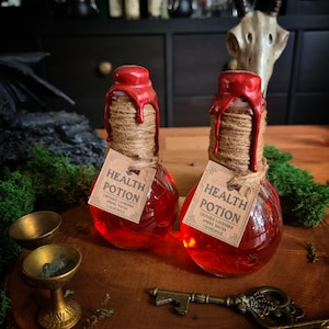 Może przedstawiać: Dwie okrągłe szklane butelki wypełnione żywym czerwonym płynem, z etykietą "Health Potion". Butelki są uszczelnione czerwoną woskiem i sznurkiem. Drewniana taca zawiera butelki, a także mosiężny klucz, różdżkę i małe mosiężne miski. Tło obejmuje czaszkę i ciemne półki.