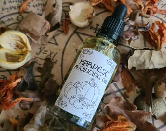 Può includere: Una bottiglia di vetro trasparente con un contagocce nero, etichettata "HARVEST anointing OIL" con un'illustrazione di zucca e mela. La bottiglia è appoggiata tra foglie secche ed elementi botanici su una superficie simile alla pergamena. Il tema generale è autunnale e naturale.