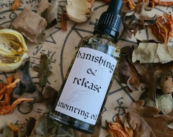Può includere: Una bottiglia di vetro trasparente con contagocce nero, etichettata "banishing & release anointing oil". La bottiglia è circondata da foglie e fiori secchi nei toni del marrone e dell'arancione, su una superficie simile alla pergamena.