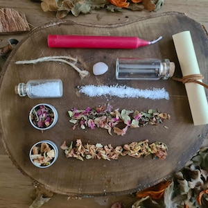May include: A collection of ritual items arranged on a wooden surface. Includes a red candle, small glass bottles, a scroll, a small stone, and dried herbs and flowers. The items are arranged in a visually appealing manner, suggesting a spiritual or crafting theme.
