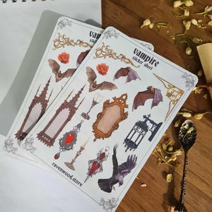 May include: Two sticker sheets with a vampire theme. The sheets feature bats, roses, gothic architecture, mirrors, and a raven. The text "vampire sticker sheet" and "ravenwood.store" are visible on the sheets. The stickers are on a wooden surface.
