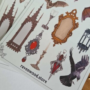 May include: Sheets of stickers featuring gothic and Victorian-inspired illustrations. Designs include bats, roses, ornate mirrors, jewelry, and architectural elements. The text "ravenwood.store" is printed on the stickers.